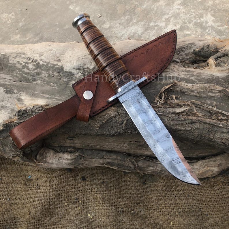 Może przedstawiać: N&oacute;ż myśliwski ze stali damasceńskiej z brązową sk&oacute;rzaną rękojeścią i pochwą. Ostrze ma unikalny wz&oacute;r, a rękojeść wykonana jest ze sk&oacute;rzanych warstw. Pochwa jest zabezpieczona metalowym zatrzaskiem. N&oacute;ż spoczywa na kawałku drewna.