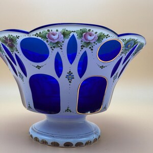 Op de afbeelding: Een decoratieve glazen kom met een witte buitenkant en levendige blauwe accenten. De kom heeft handgeschilderde roze rozen en groen blad. De basis is wit met gouden details. Een uniek pronkstuk.