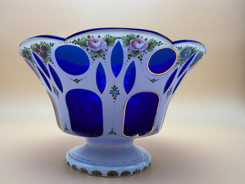 Op de afbeelding: Een decoratieve glazen kom met een witte buitenkant en levendige blauwe accenten. De kom heeft handgeschilderde roze rozen en groen blad. De basis is wit met gouden details. Een uniek pronkstuk.