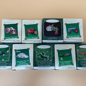 May include: A collection of ten miniature Christmas ornaments featuring various vehicles, including antique tractors, a steam locomotive, a car carrier, and a classic car. The ornaments are packaged in individual boxes with a red and green color scheme and the word "Keepsake" printed on each box.