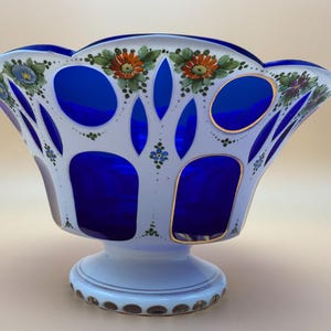 Op de afbeelding: Een decoratieve glazen kom met een wit en blauw ontwerp. De kom heeft een geschulpte rand en is versierd met handgeschilderde bloemmotieven in oranje, blauw en groen. De kom heeft een voet en gouden accenten.