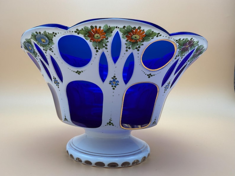 Op de afbeelding: Een decoratieve glazen kom met een wit en blauw ontwerp. De kom heeft een geschulpte rand en is versierd met handgeschilderde bloemmotieven in oranje, blauw en groen. De kom heeft een voet en gouden accenten.