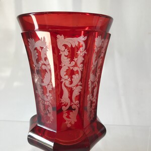 May include: A red glass vase with a white etched floral design. The vase has a hexagonal base and a flared top.