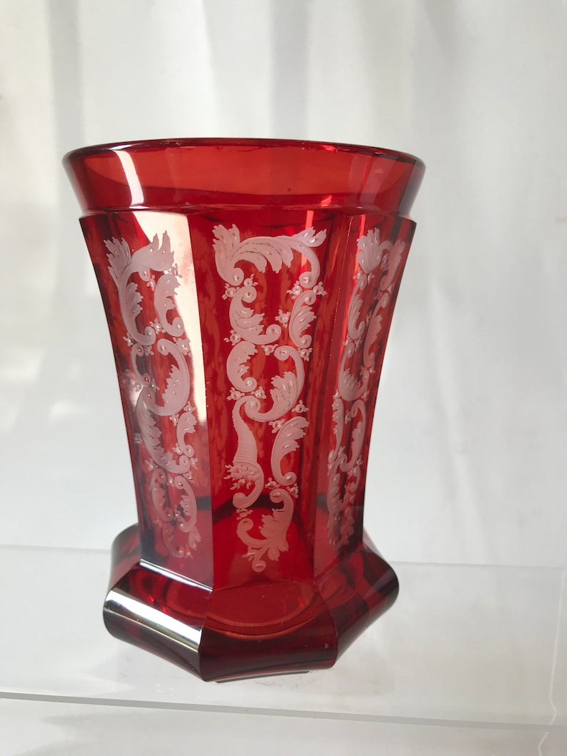 May include: A red glass vase with a white etched floral design. The vase has a hexagonal base and a flared top.