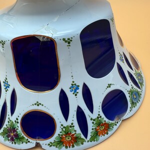 Op de afbeelding: Een witte keramische voetbakschaal met een blauw interieur en een decoratief uitgesneden ontwerp. De schaal heeft handgeschilderde bloemaccenten in oranje, paars en blauw. De schaal heeft een scheur aan de zijkant.