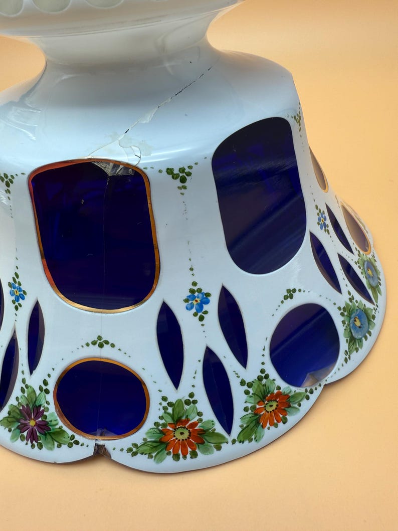 Op de afbeelding: Een witte keramische voetbakschaal met een blauw interieur en een decoratief uitgesneden ontwerp. De schaal heeft handgeschilderde bloemaccenten in oranje, paars en blauw. De schaal heeft een scheur aan de zijkant.