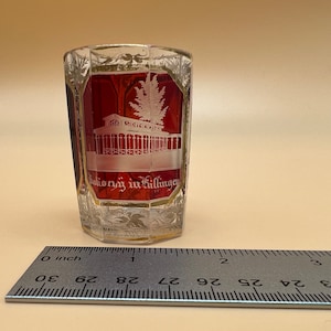 Puede incluir: Un pequeño vaso de chupito de vidrio rojo con un diseño grabado en blanco de un edificio y un árbol. El vaso tiene un borde dorado y el texto "Salioruny in Killingen" está grabado en la parte inferior.