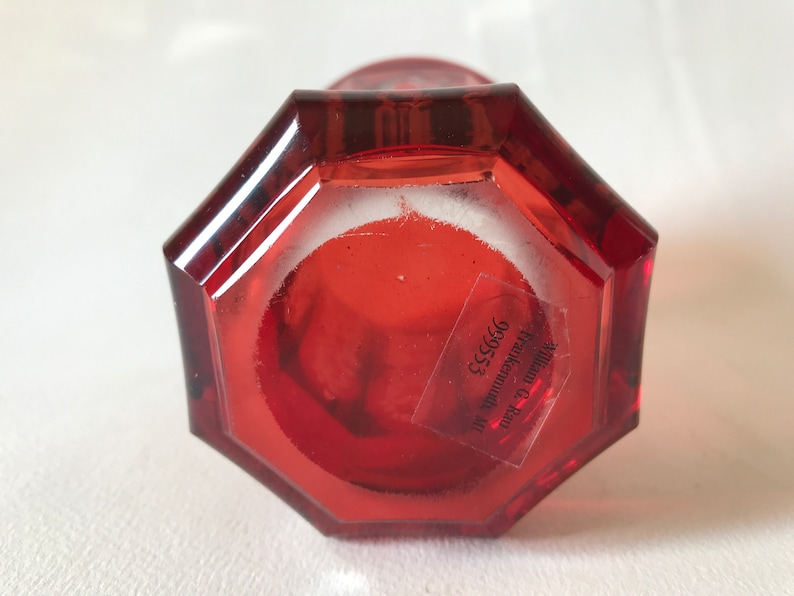 May include: Red glass vase with an octagonal base. The bottom of the vase has a label that reads "William C. Kraus Frankenmuth, MI 99653".