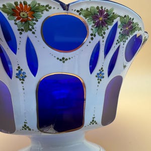 Op de afbeelding: Een witte en blauwe glazen kom met een voet. De kom heeft blauwe uitsparingen en is versierd met handgeschilderde bloemen in oranje, paars en blauw. Gouden randen accentueren de blauwe secties. De kom heeft een geschulpte rand.
