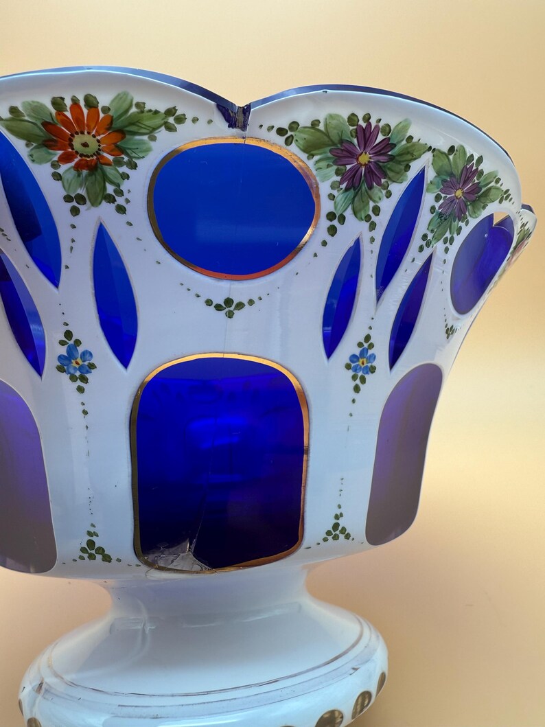 Op de afbeelding: Een witte en blauwe glazen kom met een voet. De kom heeft blauwe uitsparingen en is versierd met handgeschilderde bloemen in oranje, paars en blauw. Gouden randen accentueren de blauwe secties. De kom heeft een geschulpte rand.
