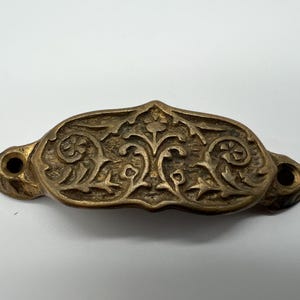 Puede incluir: Un tirador de cajón de latón con un diseño floral ornamentado. El tirador tiene dos agujeros para el montaje.