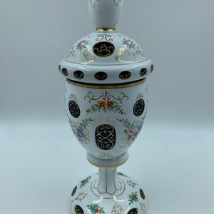 May include: White ceramic vase with a gold and black floral design. The vase has a round body and a tall, slender neck. The vase is decorated with a floral pattern and gold accents.