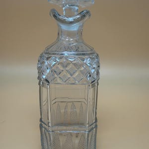 May include: A clear glass decanter featuring a square base and a neck with a flared rim. The upper body displays a diamond-cut pattern, while the lower section is decorated with vertical, leaf-shaped panels containing a fine cross-hatched texture.