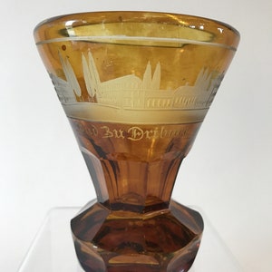 May include: A brown glass goblet with an etched design of a building and trees. The goblet has a flared top and a faceted base. The text "Bud Bud Dribnar" is etched on the side of the goblet.