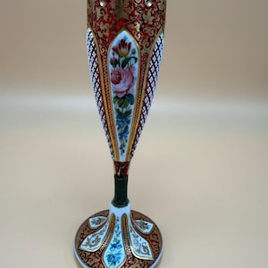 Op de afbeelding: Een hoge, slanke glazen vaas met een donkerrood en wit ruitpatroon, geaccentueerd met ingewikkeld goudfiligraan. Het centrale paneel toont een handgeschilderd bloemstuk van roze rozen en blauwe bloemen, geplaatst op een uitlopende voet.