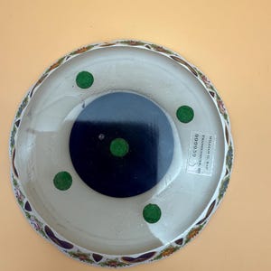 Op de afbeelding: Een witte decoratieve schaal met een bloemenrand. De schaal heeft een donkerblauwe kern en groene viltvoetjes. De onderkant van de schaal heeft een sticker met tekst.