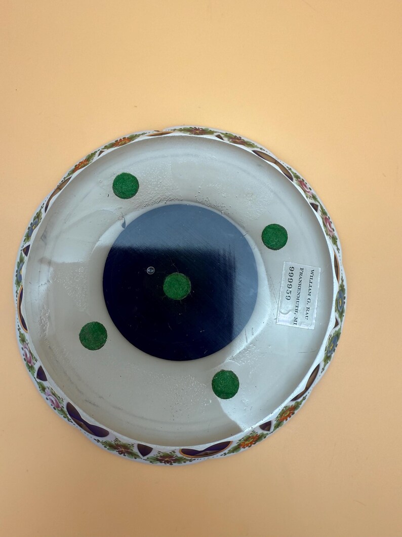 Op de afbeelding: Een witte decoratieve schaal met een bloemenrand. De schaal heeft een donkerblauwe kern en groene viltvoetjes. De onderkant van de schaal heeft een sticker met tekst.