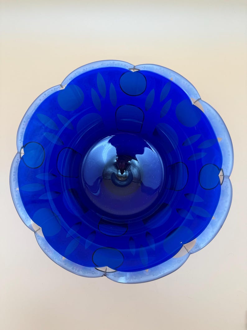 Op de afbeelding: Een levendige blauwe glazen kom met een geschulpte rand. De kom heeft een bloemmotief met donkerblauwe cirkels en bladvormen. De rand is een lichtere tint blauw, wat een contrasterende rand cre&euml;ert. De binnenkant van de kom reflecteert het licht.