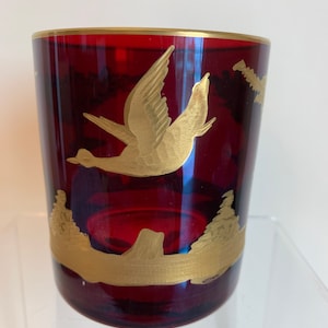 Puede incluir: Un vaso rojo de vidrio con un diseño de pájaro dorado y detalles dorados. El pájaro está volando en el aire y hay árboles en el fondo.