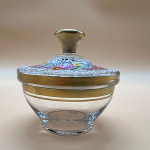 Könnte beinhalten: Ein klares Glasgefäß mit einem goldenen Deckel. Der Deckel ist mit einem Blumenmuster in Rosa-, Blau- und Gelbtönen verziert. Das Gefäß hat einen goldenen Streifen um die Mitte.