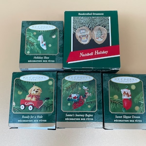 Könnte beinhalten: Fünf Weihnachtsbaumschmuck-Boxen mit verschiedenen Designs. Die Boxen sind grün und weiß mit dem Schriftzug "Keepsake Ornament" auf der Vorderseite. Die Ornamente sind ein Fest-Schuh, ein Nussschalen-Weihnachten, ein Welpe in einem roten Wagen, Santas Reise beginnt und eine Katze in einem Strumpf.