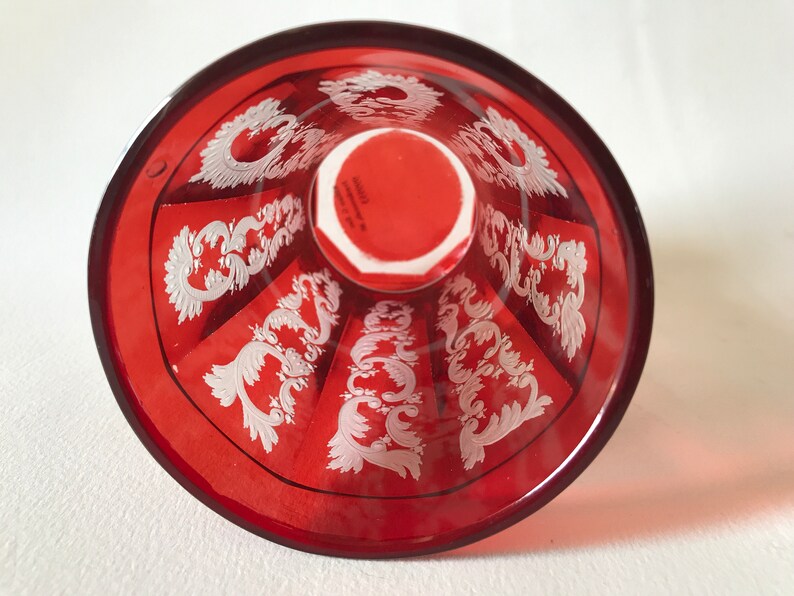 May include: The bottom of a red glass goblet with a white etched design. The design features a repeating pattern of stylized floral motifs.