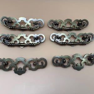 May include: A set of six antique-style brass drawer pulls with ornate, scrollwork backplates. Four of the pulls feature matching swinging handles, while two backplates are shown without handles, displaying the mounting points clearly.