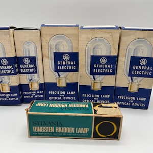 May include: Five vintage General Electric precision lamps for optical devices in their original boxes. The boxes are blue and white with the General Electric logo and text. A Sylvania Tungsten Halogen Lamp box is in the foreground.