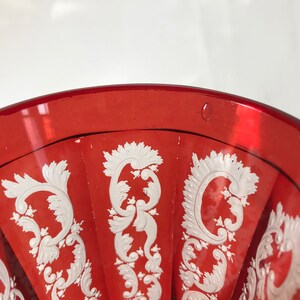 May include: A red glass goblet with a white, intricate, floral design. The goblet has a wide, round bowl and a short stem.