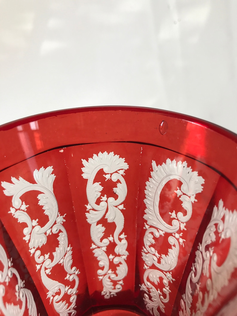 May include: A red glass goblet with a white, intricate, floral design. The goblet has a wide, round bowl and a short stem.