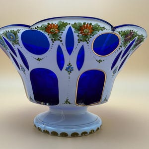 Op de afbeelding: Een decoratieve glazen kom met een witte buitenkant en een levendig blauwe binnenkant. De kom heeft een geschulpte rand en is versierd met handgeschilderde bloemmotieven in oranje, groen en blauw. De basis is lichtblauw met gouden accenten.