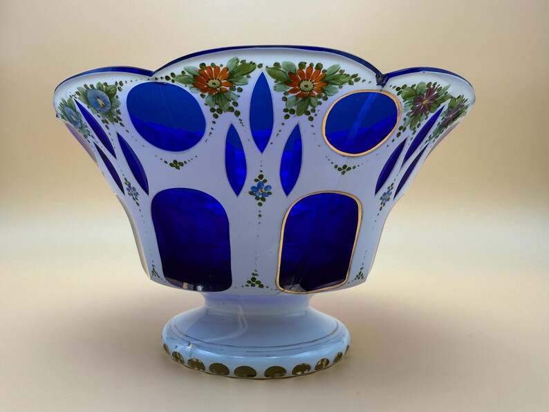 Op de afbeelding: Een decoratieve glazen kom met een witte buitenkant en een levendig blauwe binnenkant. De kom heeft een geschulpte rand en is versierd met handgeschilderde bloemmotieven in oranje, groen en blauw. De basis is lichtblauw met gouden accenten.