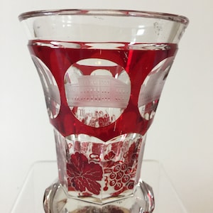 May include: A clear glass drinking glass with a red, ruby-colored design. The glass has a wide, flared mouth and a round base. The design features a white etched image of a building and a grapevine with grapes.