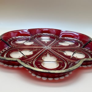 May include: A red glass serving tray with a white and gold design. The tray has a scalloped edge and a beaded rim.