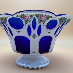 Op de afbeelding: Een decoratieve glazen kom met een witte buitenkant en een levendig blauwe binnenkant. De kom heeft handgeschilderde roze rozen en groene bladeren rond de rand. De basis is wit met een gouden rand. Een uniek stuk voor woondecoratie.