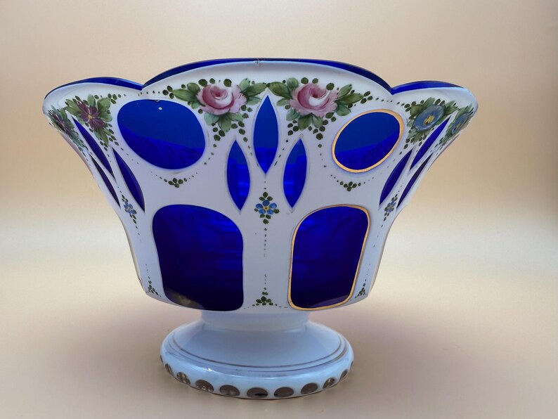 Op de afbeelding: Een decoratieve glazen kom met een witte buitenkant en een levendig blauwe binnenkant. De kom heeft handgeschilderde roze rozen en groene bladeren rond de rand. De basis is wit met een gouden rand. Een uniek stuk voor woondecoratie.