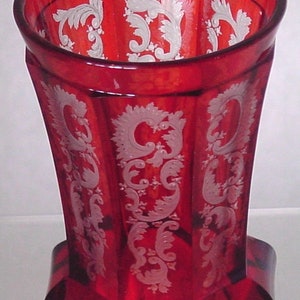 Peut inclure: Un vase en verre rouge avec un motif floral gravé en blanc. Le vase a une forme hexagonale et une large ouverture.
