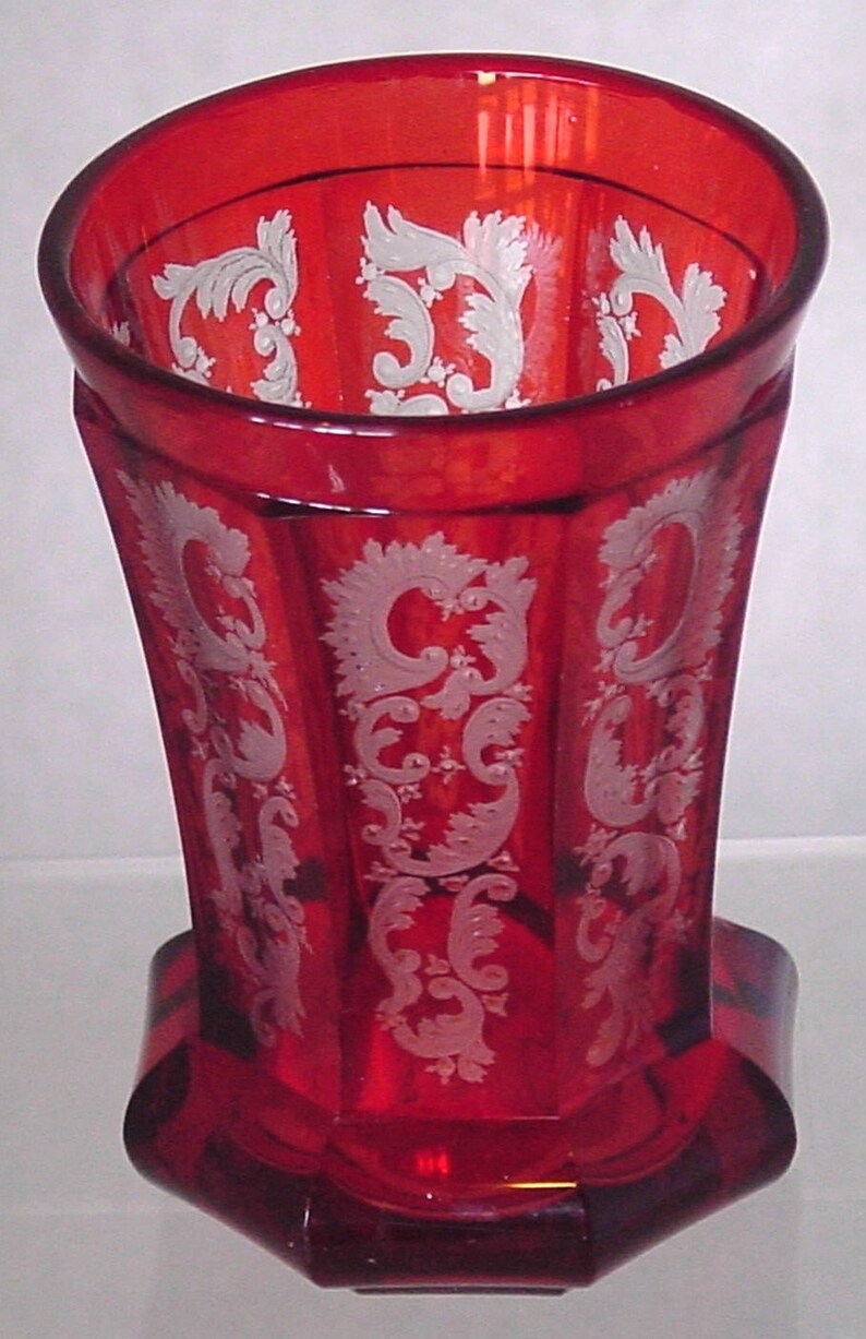 May include: A red glass vase with a white etched floral design. The vase has a hexagonal shape and a wide mouth.