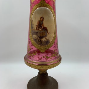 Op de afbeelding: Een roze glazen vaas met gouden accenten en een geschilderd tafereel van een persoon die vist. De vaas heeft een houten voet.