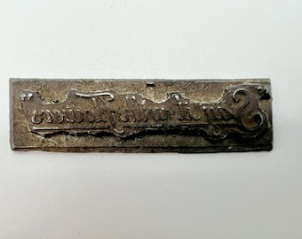May include: A rectangular, antique metal stamp with the cursive text "casual following". The stamp has a weathered, textured appearance, with the letters slightly raised. The edges of the stamp are also textured.