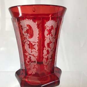 May include: A red glass vase with a white etched floral design. The vase has a hexagonal base and a flared top.