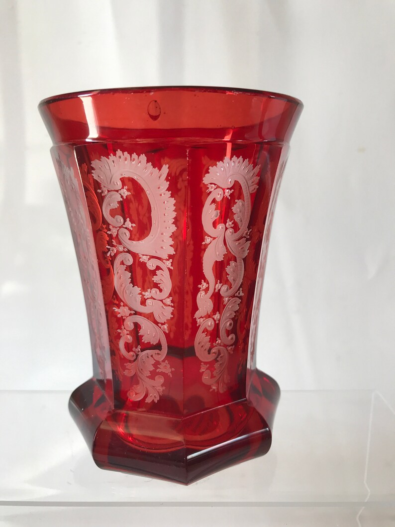 May include: A red glass vase with a white etched floral design. The vase has a hexagonal base and a flared top.