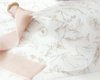 May include: White tissue paper with a delicate floral pattern in brown ink. The paper is folded in half and a pale pink satin ribbon is draped over it.