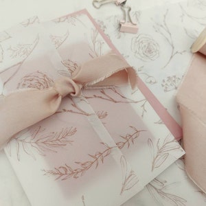 May include: A blush-coloured ribbon tied around a floral-patterned envelope. The envelope is made of translucent paper with a delicate botanical design. A spool of matching ribbon and a paper clip are also visible.