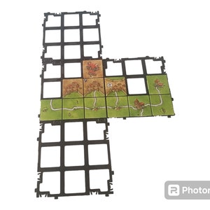 Può includere: Un gioco da tavolo con un tabellone a griglia nero e una varietà di tessere colorate che rappresentano una scena di villaggio medievale. Le tessere sono disposte a forma di L sul tabellone.