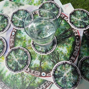 May include: A set of 10 round coasters with a forest scene and the text "The Edge Shaman Designs" printed on each coaster. The coasters are arranged in a circle around a glass of water.