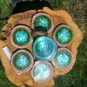 May include: A set of six wooden coasters with a painted forest scene in shades of green and blue, arranged around a clear glass. The coasters are made from wood slices with natural bark edges. The items are displayed on a large wooden slab.