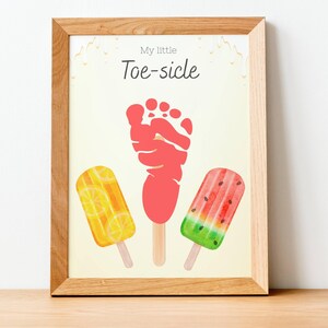May include: A framed print featuring a red footprint resembling a popsicle, flanked by watercolor illustrations of lemon and watermelon popsicles. The text "My little Toe-sicle" is at the top. The frame is made of light-colored wood.