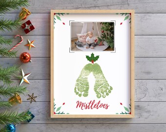 May include: A white frame with a green and red holly design. The frame has a photo of a child in a white onesie standing in front of a Christmas tree. The frame also has the words "Mistletoes" printed on it.