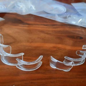 May include: Clear plastic dental retainers are arranged on a wooden surface. Several retainers are visible, some in individual plastic bags. The retainers are curved and designed to fit the shape of teeth. The image shows the product in a clear, transparent color.